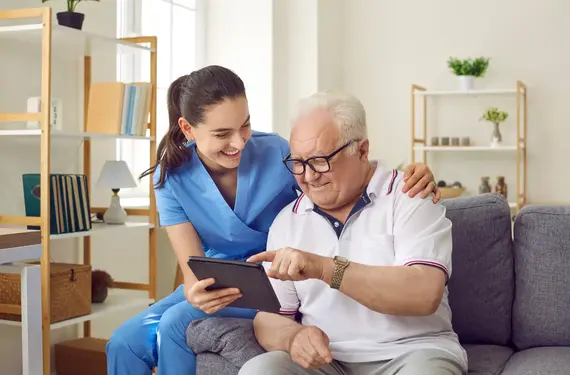 A smiling home caregiver in blue scrubs sits beside an elderly man on a sofa, helping him navigate a tablet computer in a bright living room.