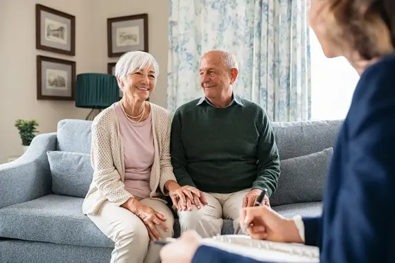 A smiling elderly couple sit on a sofa during a home care for elderly consultation, as a professional takes notes opposite them.