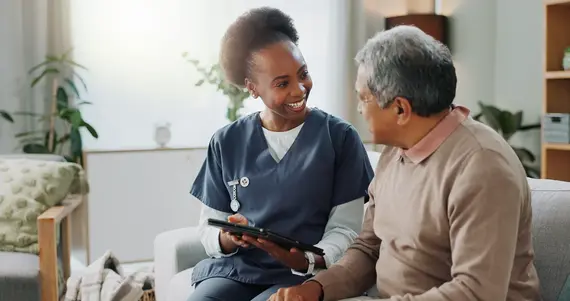 A home carer in navy scrubs smiles warmly at an elderly man whilst sharing information on a tablet during a visit at his home.