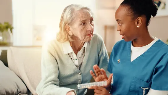 A homecare nursing professional in blue scrubs explains a weekly pill organiser to an elderly woman in her living room.