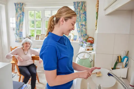 A cheerful carers home visit shows a young woman in a blue polo shirt washing up whilst an elderly lady relaxes at the kitchen table.