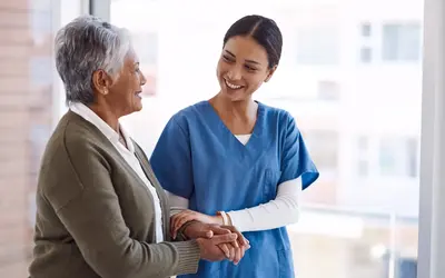 An elderly caregiver in blue scrubs smiles warmly whilst holding the hands of an older woman in a bright care setting.