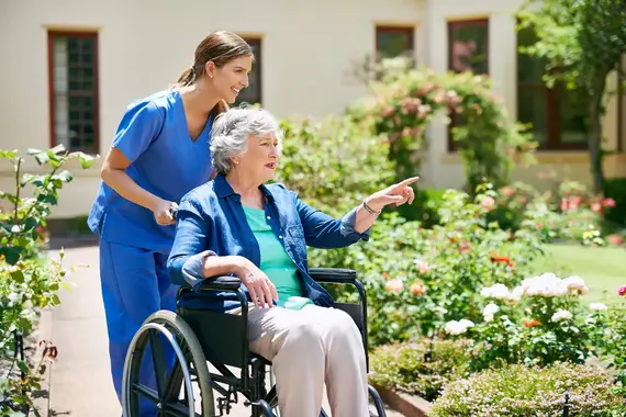 An elderly caregiver in blue scrubs pushes a smiling older woman in a wheelchair through a sunny rose garden outside a care home.