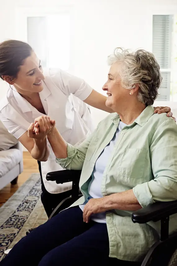 A caring professional in a white uniform holds hands with a smiling elderly woman in a wheelchair, showing warm carers for elderly at home support.