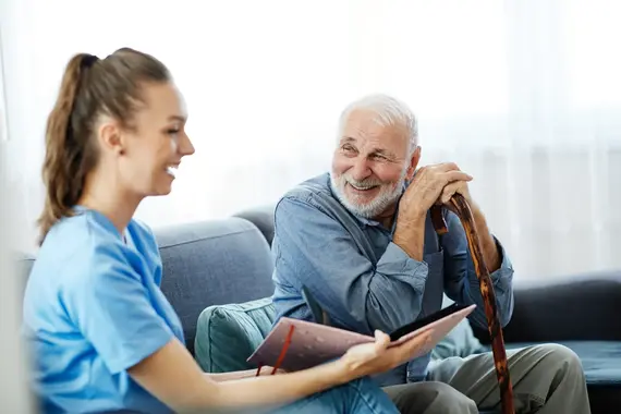 A carers home visit showing a young female carer in blue scrubs chatting warmly with a smiling elderly man holding a walking cane.