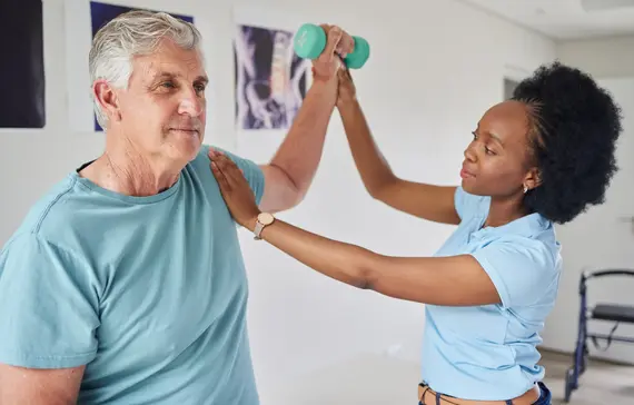A senior caregiver supports an older man as he raises a small dumbbell overhead during a shoulder rehabilitation session in a clinical room.