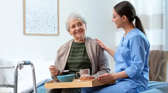 A carers home visit showing a young carer in scrubs offering a breakfast tray to a smiling elderly woman seated on her bed.