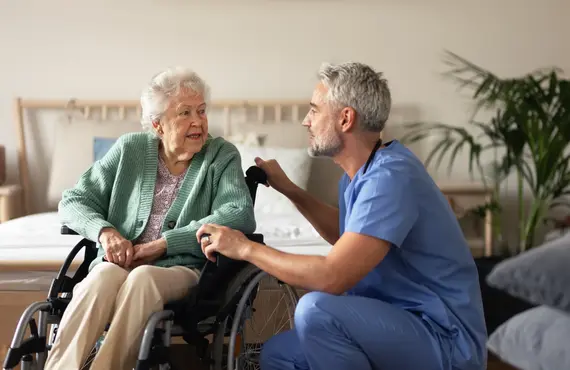 A male carer in blue scrubs kneels attentively beside an elderly woman in a wheelchair at a carers home, speaking with her warmly.