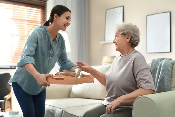 A smiling home carer for elderly hands a wooden breakfast tray to an older woman seated comfortably on a sofa in a bright living room.