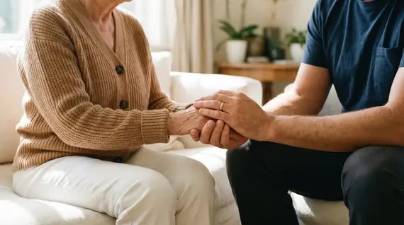 A private carer sits beside an elderly woman on a sofa, tenderly holding her hands in a warm, sunlit living room.