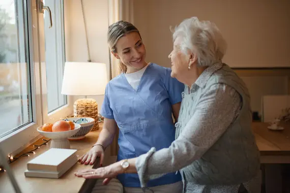 A carers home visit showing a smiling carer in blue scrubs sharing a warm moment with an elderly woman by a sunlit window.