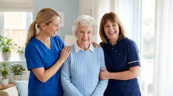 Two smiling carers in blue uniforms provide home care services to a happy elderly woman in a bright, welcoming room.