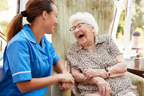 A smiling carer in a blue uniform holds hands with a laughing elderly woman during a carers home visit.