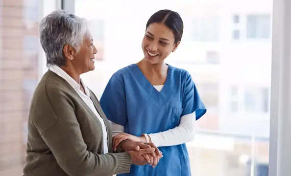 An elderly caregiver in blue scrubs smiles warmly whilst holding the hands of an older woman in a bright care setting.