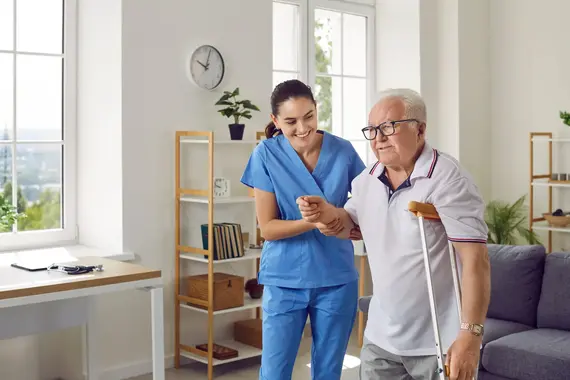 A nurse in blue scrubs provides in home care by steadying an elderly man using a crutch as he walks across a bright living room.
