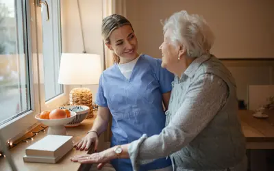 A carers home visit showing a smiling carer in blue scrubs sharing a warm moment with an elderly woman by a sunlit window.