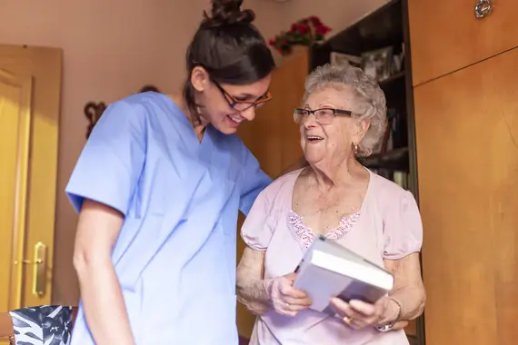 A smiling carer in blue scrubs shares a joyful moment with an elderly woman during a carers home visit, as the woman holds a book and laughs.