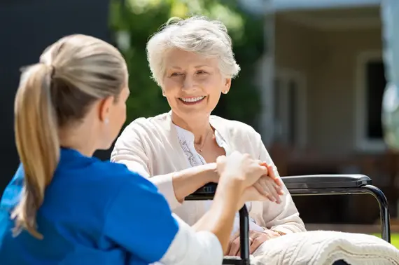 An elderly caregiver in a blue uniform holds the hands of a smiling older woman seated in a wheelchair outside.