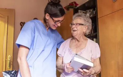 A smiling carer in blue scrubs shares a joyful moment with an elderly woman during a carers home visit, as the woman holds a book and laughs.