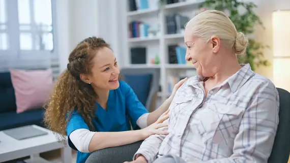 A young carer in blue scrubs smiles and places a reassuring hand on an elderly woman's shoulder while caring for the elderly at home.