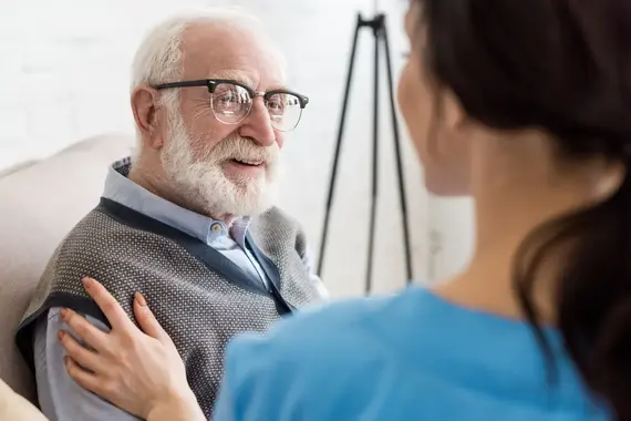 A carers home visit in action as a female carer in blue scrubs gently places her hand on a smiling elderly man's shoulder.