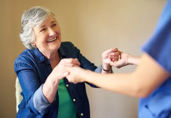 A cheerful elderly woman smiles broadly as an elderly caregiver at home holds both her hands in a caring, supportive gesture.