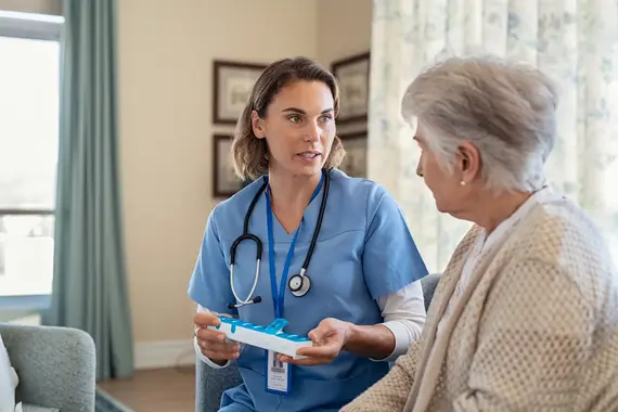 A nurse in blue scrubs holds a weekly pill organiser while providing home care services to an elderly woman in her living room.