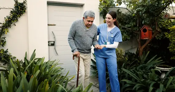 Dementia care worker supporting an older man with a walking stick in his garden at home.