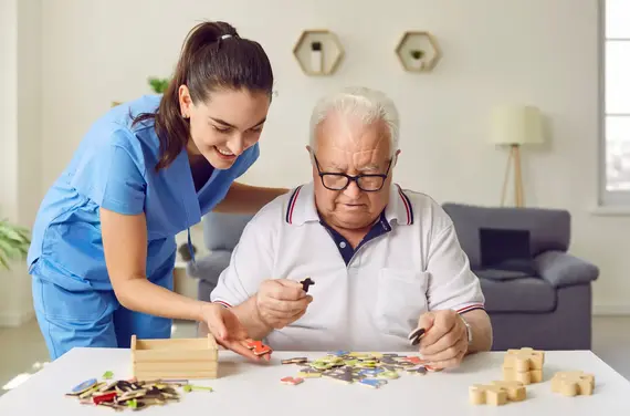 An elderly caregiver in blue scrubs smiles as she helps an older man with glasses sort wooden puzzle pieces at a home care table.
