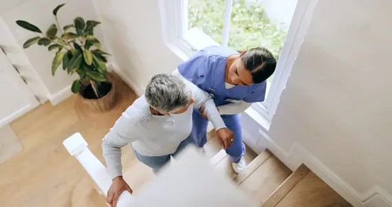 A carers home visit showing a nurse in blue scrubs carefully supporting an elderly person up the stairs inside a bright home.