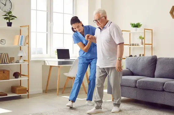 A carers home visit showing a nurse in blue scrubs gently supporting an elderly man as he walks across a sunlit living room.