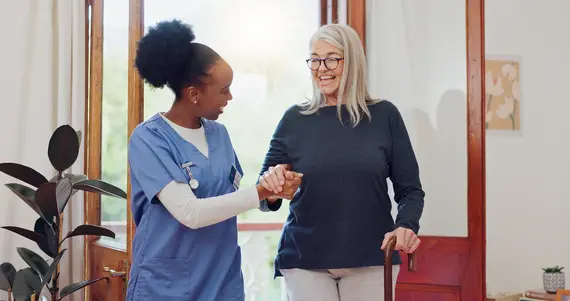 A home carer in blue scrubs holds the hand of a smiling older woman using a walking cane inside a bright, welcoming home.