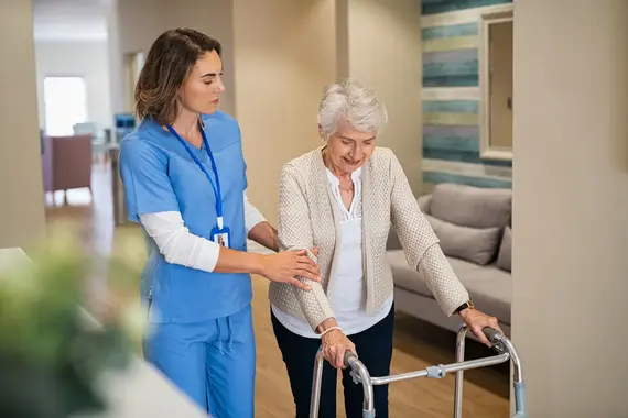 An elderly caregiver in blue scrubs steadies a smiling older woman using a walking frame in a bright care home corridor.