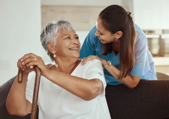 A smiling carers home visit shows a young carer in blue scrubs placing a reassuring hand on an elderly woman's shoulder as they share a warm moment.