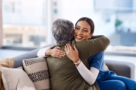 A smiling carers home professional in blue scrubs warmly embraces an elderly client on a sofa during a home visit.