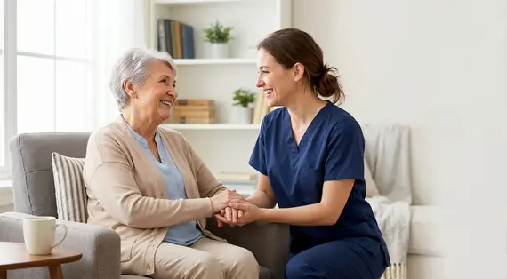 A homecare worker in navy scrubs smiles warmly whilst holding the hands of an elderly woman seated on a sofa in a light-filled living room.